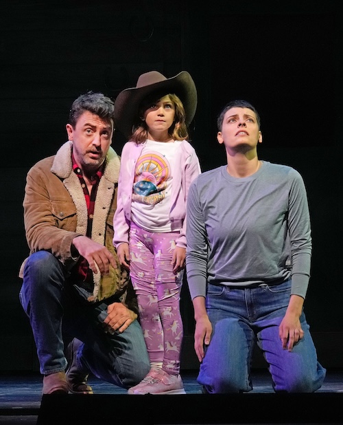 A man, a young child wearing a cowboy hat, and a woman kneel together on a stage, all looking upward with concerned or awed expressions. The child wears pink pajamas with a rainbow graphic, while the adults are dressed casually. The scene is dimly lit, suggesting a theatrical performance.