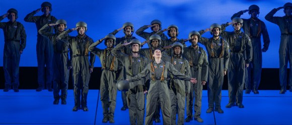 A group of performers dressed in matching green flight suits and helmets stand in formation on a stage lit in blue. Most of them are saluting with serious expressions, while one person at the front center stands with arms outstretched, head tilted back, and mouth open as if singing or shouting. The group is arranged in rows, creating a symmetrical, choreographed scene against a minimalist blue backdrop.
