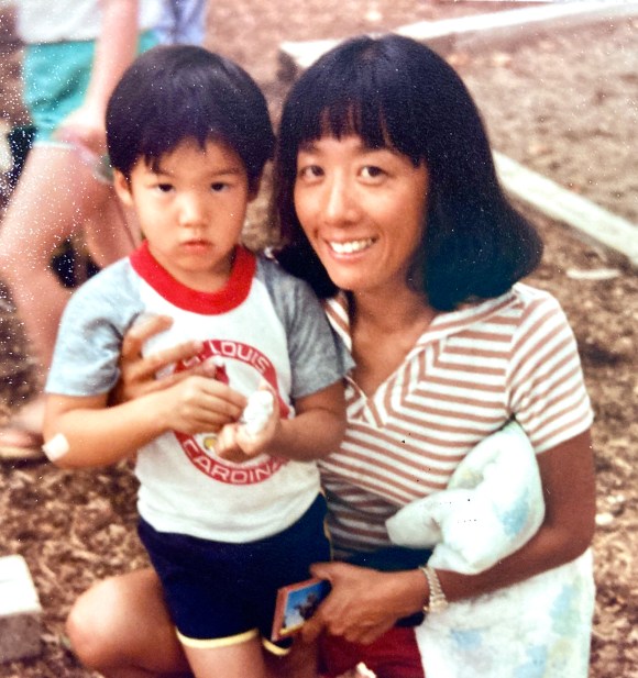 A slightly faded, vintage-looking photo of a smiling woman with short dark hair kneeling beside a young child outdoors. The child, wearing a light-colored T-shirt with red trim and dark shorts, looks serious while holding a small object. The woman, wearing a striped shirt, has her arm around the child and holds a soft item in her other arm. The background shows a dirt ground with scattered leaves and blurred figures, suggesting a casual outdoor setting.
